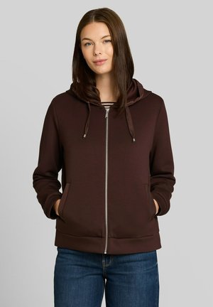 Frau mit glatten braunen Haaren trägt einen braunen Hoodie mit Reißverschluss, die Hände in den Taschen und blaue Jeans vor grauem Hintergrund.