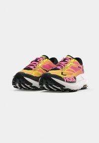 Une paire de chaussures de course athlétiques jaunes et roses avec des semelles noires et des lacets roses, inclinées côte à côte sur un fond blanc.
