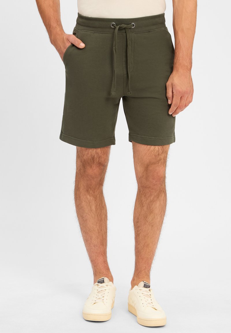Dunkelgrüne Baumwollmischung-Shorts mit einem Kordelzugbund, seitlichen Taschen und umgeschlagenen Säumen, kombiniert mit beigen Sneakers.