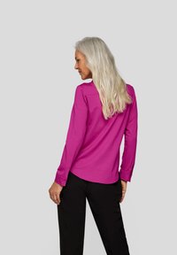 Fuchsia long-sleeve shirt with a rounded hem and button accents, paired with black pants. Smooth texture, simple design.
