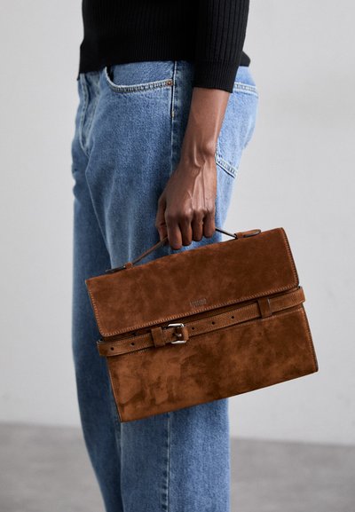 Geantă din ante maro, cu un design plat, mânere mari și o curea cu o cataramă argintie. Purtați cu blugi bleu de denim.