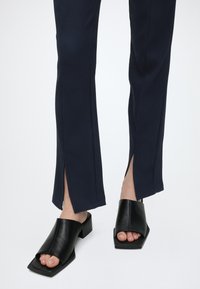 Jambes portant un pantalon côtelé bleu marine avec des fentes à l'avant, associé à des sandales mules noires à talon carré et à bout ouvert sur fond blanc.