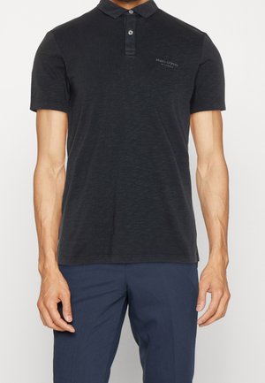 Man die een zwart poloshirt met korte mouwen en een klein logo op de borst draagt, gecombineerd met donkerblauwe broek, staand tegen een witte achtergrond.