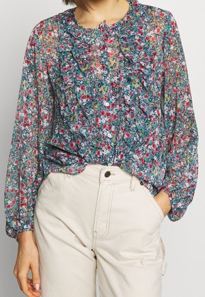 Femme portant un chemisier transparent à manches longues à motifs floraux avec des volants et un pantalon blanc taille haute, une main dans la poche, sur un fond uni.