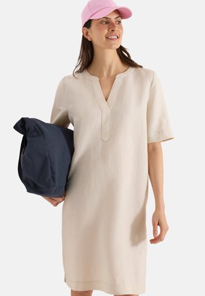 Femme portant une robe beige à manches courtes et une casquette rose, tenant une veste bleu marine foncé pliée dans sa main gauche, regardant vers la droite, souriante.