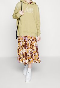 Olivgrön hoodie med logotyp, ihopparad med en mönstrad, plisserad midi-kjol i bruna, lila och krämfärgade nyanser. Vita sneakers fulländar looken.