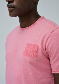 Camiseta rosa de manga corta en tejido suave, con un logo texturizado de "SALSA JEANS" en el pecho. Cuello redondo con un ligero brillo.