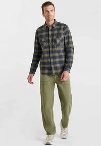Chemise à carreaux bleu marine et vert olive à manches longues avec poches poitrine, associée à un pantalon cargo vert olive et des baskets grises.