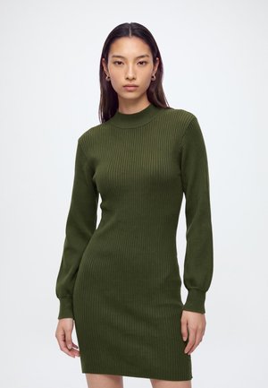 Femme aux longs cheveux foncés portant une robe en maille côtelée olive ajustée, à manches longues, et des boucles d'oreilles anneaux dorées.