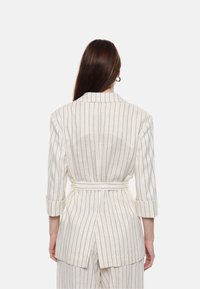 Blazer a righe in beige chiaro e blu navy, realizzato in lino, presenta una vita con cintura, maniche a tre quarti e un dettaglio con spacco posteriore.