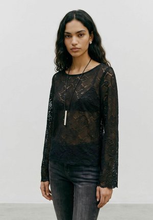 Femme aux cheveux noirs ondulés porte un haut à manches longues en dentelle noire par-dessus un soutien-gorge noir, un jean foncé, des boucles d'oreilles argentées et un long collier pendentif.