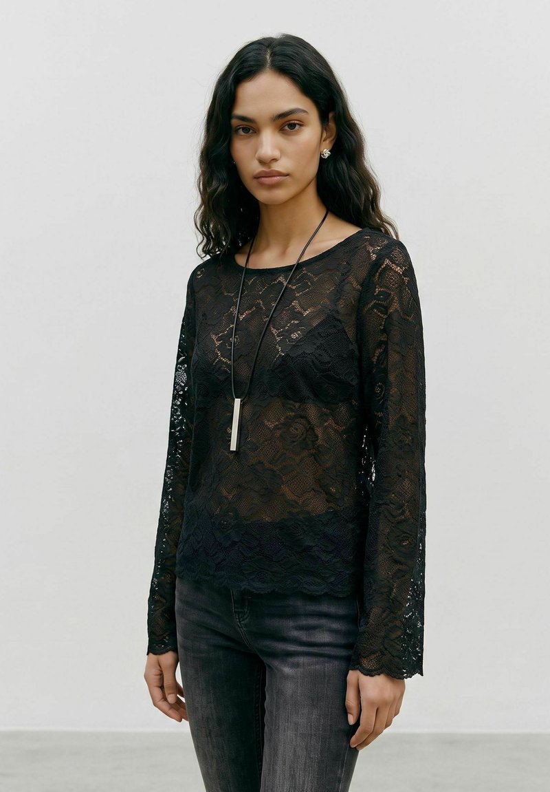 Femme aux cheveux noirs ondulés porte un haut à manches longues en dentelle noire par-dessus un soutien-gorge noir, un jean foncé, des boucles d'oreilles argentées et un long collier pendentif.