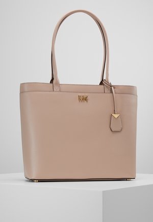 Sac cabas en cuir beige avec doubles poignées, logo MK doré et étiquette en cuir suspendue, posé sur un socle blanc devant un fond gris.