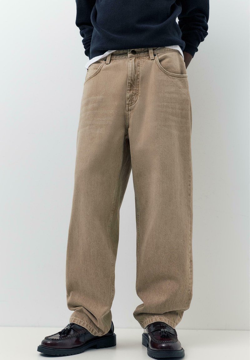 Persoon die losse beige broek, zwarte loafers met kwastjes en een donker sweatshirt draagt, met de handen in de zakken.