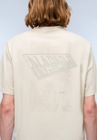 Lyscremet t-shirt med en struktureret, rektangulær grafik, der fremhæver ordet "NAPAPIJRI" og året "1987" i et diskret design.