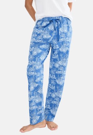 Osoba ubrana w luźne niebieskie spodnie w biały kwiatowy wzór z regulowanym paskiem w talii, boso, częściowo widoczna biała bluzka.