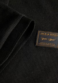 Tissu noir texturé avec un revers roulé et une étiquette Jack & Jones indiquant "Superior Apparel" cousue sur la surface.