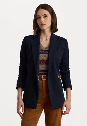 Donna che indossa un blazer blu navy sopra un maglione a righe in maglia, pantaloni in velluto a coste marroni e una cintura marrone, in piedi davanti a uno sfondo semplice.