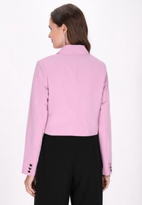 Blazer rosa corto con maniche lunghe, colletto a scollo a V e dettagli in bottone nero sui polsi. Tessuto liscio con una vestibilità su misura e cuciture sul retro.