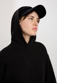 Czarna bluza z kapturem z delikatnie wyhaftowanym logo w ciemnym kolorze. Noszona z czapką z kordurowego materiału w czarnym kolorze z lekko zakrzywionym daszkiem.