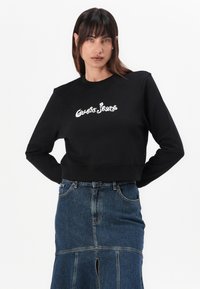 Sweatshirt cropped preto com o logotipo "Guess Jeans" em branco. Possui mangas longas e um decote redondo, combinado com uma saia de ganga.