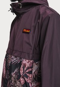 Närbild av en lila huvan jacka med en blomsterprydd nedre del och ett rödgult Scotch & Soda-logotypmärke på bröstet.