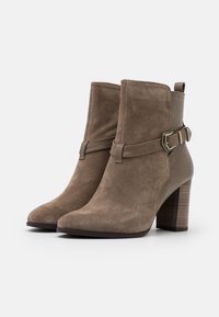 Stivaletti alla caviglia in suede marrone con punta affusolata, tacco a blocco e una fascia decorativa con dettagli a fibbia in metallo. Texture liscia e design minimalista.