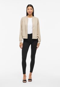 Beige bomberjakke med frontlukking, ribbestrikkede mansjetter og en avslappet passform. Kombinert med svarte leggings og svarte ankelstøvletter med åpen tå.