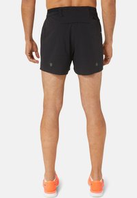 Black athletic shorts with a zippered back pocket and rounded hems. Smooth fabric texture, paired with bright orange running shoes.
