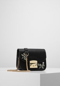 Černá kožená crossbody taška se zlatým řetízkem, zlatým zapínáním a bílou květinovou výšivkou na předním klopě, umístěná na bílém podstavci.