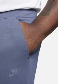 Nike sportbroek in blauwe stof, met een zijzakdetail, een gladde textuur en een subtiele logo in de onderste hoek.