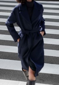 En person iklädd en mörkblå bälte kappa och grå stövlar går över ett zebramönstrat övergångsställe.