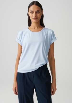 Vrouw met lang donker haar, gekleed in een lichtblauw T-shirt met korte mouwen en donkerblauwe broek, staand tegen een effen grijze achtergrond.