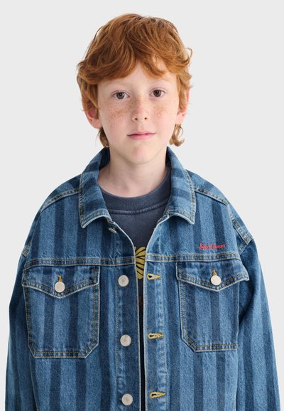Garçon aux cheveux roux et taches de rousseur portant une veste en denim rayée bleue par-dessus une chemise sombre, regardant droit devant avec une expression neutre.