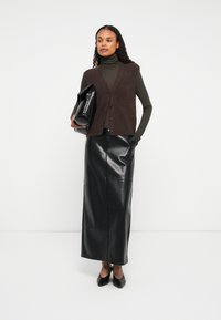 Kobieta w ciemnopomarańczowej, bezrękawnikowej kamizelce na oliwkowym golfie, czarnej skórzanej maxi spódnicy, czarnych szpilkach, trzymająca dużą czarną torebkę kopertową.