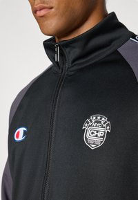 Vyras dėvi juodą striukę su užtrauktuku, pilkais peties intarpais, Champion logotipu ir NYC CHP 1919 emblemė ant krūtinės.