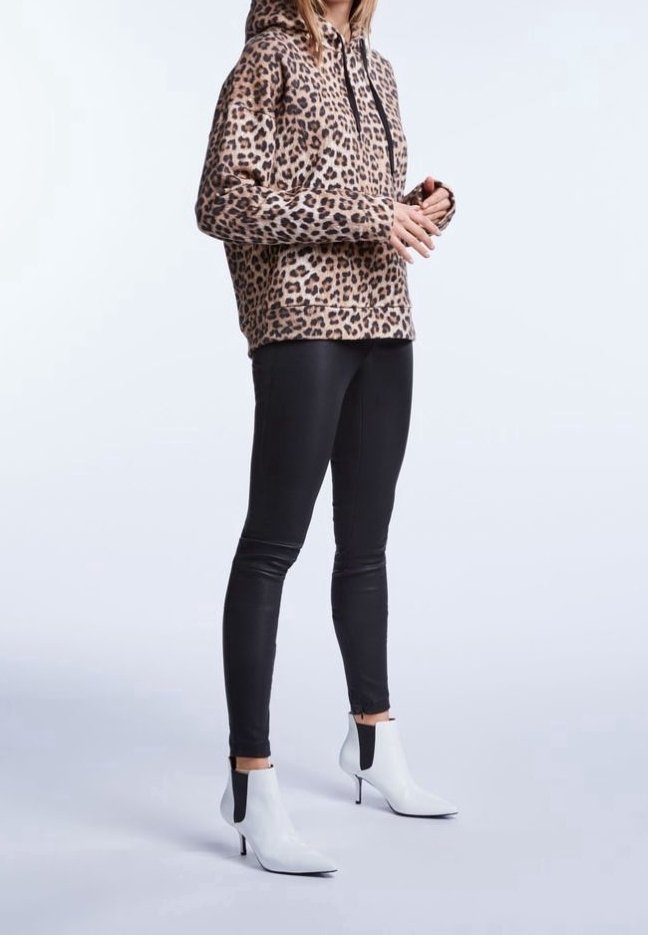Personne portant une veste à capuche imprimé léopard, un pantalon noir ajusté et des bottines blanches à talons aiguilles pointus, debout sur un fond uni.
