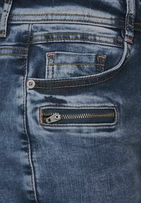 Jeans de denim azul oscuro con un bolsillo de cremallera visible, costuras amarillas en contraste y una textura ligeramente deslavada en la tela.
