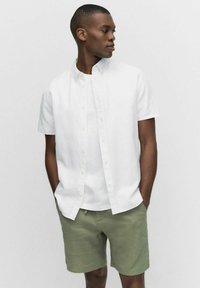 Witte shirt met korte mouwen en een button-down kraag, gedragen over een wit T-shirt en olijfgroene shorts, die een ontspannen pasvorm en lichte textuur tonen.