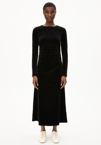 Schwarzes Samt-Kleid mit langen Ärmeln, enganliegendem Oberteil und weitem Rock. Glatte Textur, Rundhalsausschnitt und knöchellanges Design. Getragen mit hellen Schuhen.