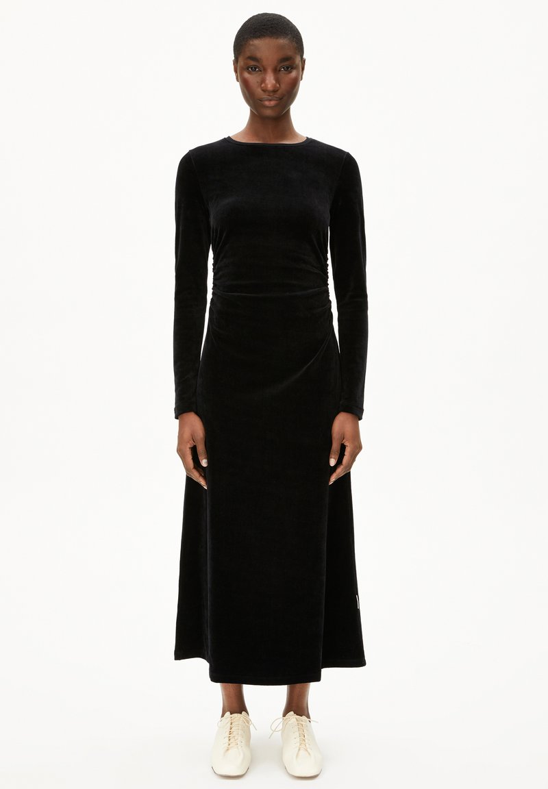 Schwarzes Samt-Kleid mit langen Ärmeln, enganliegendem Oberteil und weitem Rock. Glatte Textur, Rundhalsausschnitt und knöchellanges Design. Getragen mit hellen Schuhen.