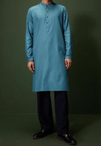 Man in een teal lange mouw kurta met zwarte broek en zwarte loafers, staand tegen een donkergroene achtergrond.