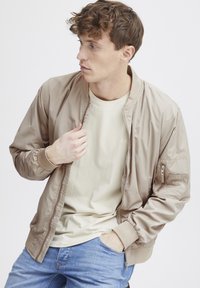 Beige bomberjakke med ribbestrikkede mansjetter og krage, glidelås foran og en lommeslipp på ermene. Brukt over en lys t-skjorte og blå jeans.