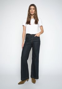 Donna in piedi che indossa una camicia bianca a maniche corte, jeans larghi scuri a vita alta e stivaletti marroni con tacco su uno sfondo bianco uniforme.
