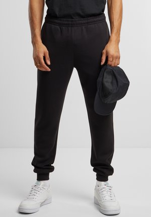 Sort træningsbukser med elastisk talje, afsluttede ben, og en glat tekstur. Matchet med hvide sneakers og en sort kasket, der holdes i hånden.