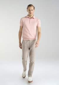 Jasnoróżowa koszulka polo z krótkim rękawem i kołnierzykiem, w połączeniu z beżowymi spodniami slim fit oraz białymi trampkami z brązowymi akcentami.