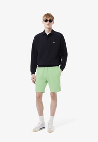 Polo-shirt à manches longues bleu marine avec un col et un logo crocodile vert, associé à un short vert clair et des baskets blanches avec des accents subtils.