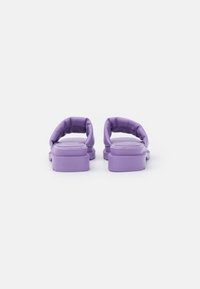Lila slip-on sandaler med en vadderad rem och en platt gummisula. Slät textur med subtila spår på sulan. Inga framträdande dekorationer.