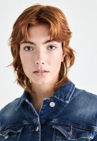 Veste en denim avec tissu bleu, fermeture à boutons sur le devant, deux poches poitrine et coutures contrastées. Le modèle a des cheveux courts, ondulés, brun-roux et porte des boucles d'oreilles créoles.