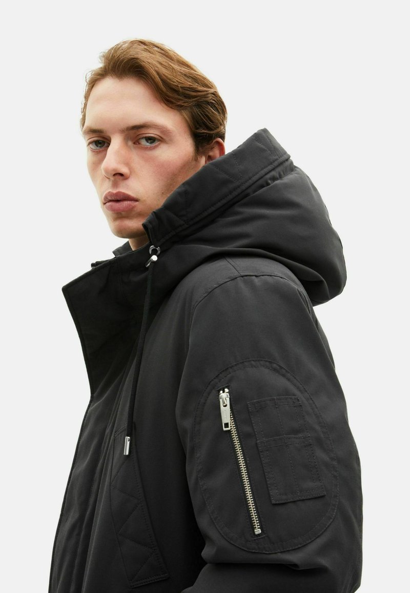 Black padded jacket with a high collar and adjustable hood. Features side zippered pocket and patch pocket on the sleeve. Smooth texture.